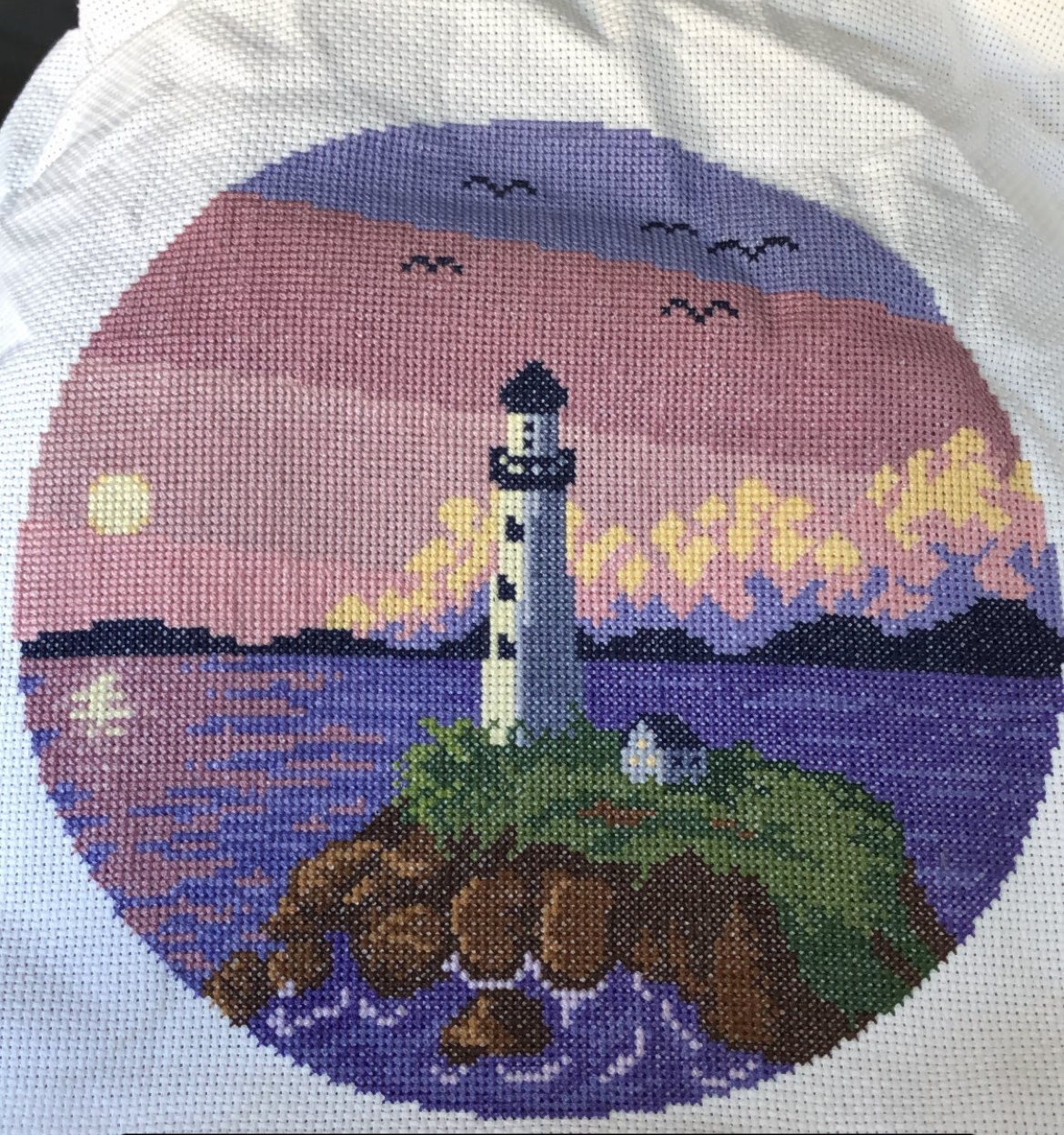 A cross stitch of a lighthouse on a small island surrounded by water and the sunset